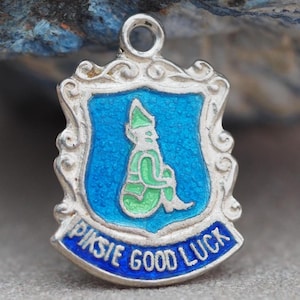 May include: A silver charm with a blue and green enamel design. The charm is shield-shaped, depicting a seated figure and the words "PIKSIE GOOD LUCK" in blue. It has a loop at the top for attaching to a chain or bracelet.