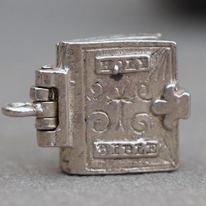 May include: Silver charm in the shape of a closed book with a cross on the cover. The book is engraved with the words "Holy Bible".