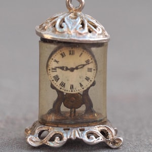 May include: A miniature silver-toned clock charm with a clear, cylindrical case. The clock face has Roman numerals and black hands. The charm has an ornate base and a decorative top with a loop for hanging.