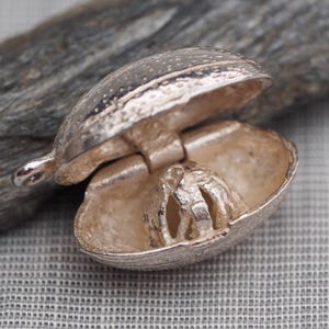 May include: A silver-toned metal locket pendant shaped like an open clam shell. The exterior has a textured, speckled surface, while the interior reveals a small, hollow, cage-like structure attached to the centre. A small loop is on the side.