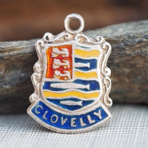 Peut inclure: Un pendentif en métal argenté en forme de blason avec des détails en émail. L'écu présente une section verticale orange avec des poissons blancs, ainsi que des bandes horizontales jaunes et bleues. Une bannière bleue en bas porte le mot CLOVELLY.