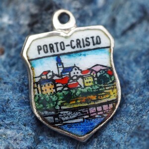 以下が含まれることがあります： 上部に「PORTO-CRISTO」の文字が入った、銀色のシールド型ペンダント。赤い屋根の建物、教会、青い空のある、海岸沿いの町のカラフルなエナメルシーンが特徴です。ペンダントの上部には、チェーンに取り付けるための小さなループが付いています。