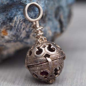 May include: A silver pendant with a filigree sphere design. The sphere has intricate cut-out patterns and a small red stone visible inside. The pendant has a loop at the top for hanging and a twisted rope-like detail above the sphere.