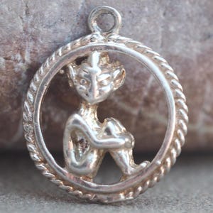 May include: A silver pendant featuring a seated fairy figure within a circular frame. The fairy is depicted with pointed ears and is holding its knees. The frame has a twisted rope design, and a loop is at the top for a chain.