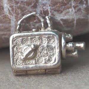 May include: A small, silver-colored charm in the shape of a vintage movie camera. The charm has a handle, lens details, and a decorative pattern on the front. The camera charm is likely made of metal and is on a gray surface.