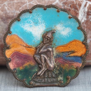 May include: A vintage brooch with a detailed design. The brooch features a silver-toned figure of a seated elf against a backdrop of a colorful landscape. The landscape includes a blue sky, orange and purple hills, and a green foreground. The brooch has a scalloped edge.