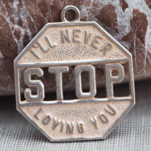 May include: An octagonal silver-toned metal pendant featuring a textured surface and raised lettering. The text reads I'LL NEVER STOP LOVING YOU, with the word STOP prominently displayed in the center. A small circular loop is attached at the top.
