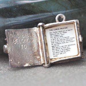 May include: A small, open silver charm in the shape of a book. The left page reads "THIS BIBLE BELONGS TO". The right page displays "THE LORDS PRAYER" in black text. A small loop is at the top for a chain.