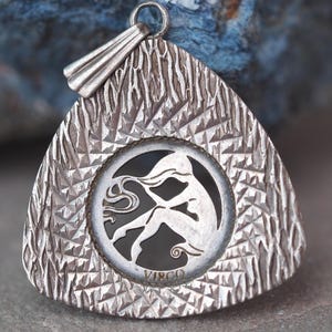May include: Silver-toned Virgo pendant with a textured triangular frame and a circular center. The center features a silhouette of a woman and the word "VIRGO". The pendant has a loop for a chain.