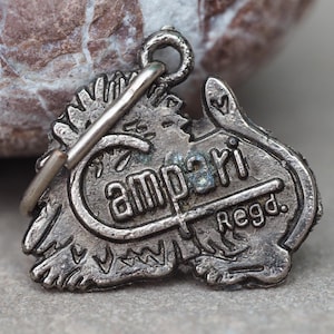 May include: A vintage silver-toned charm with the word "Campari" in a stylized font. The charm has a loop for attachment and the word "Regd." is also visible. The charm is likely a collectible or a piece of jewelry.