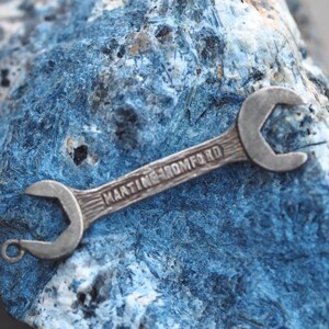 May include: A small, silver wrench with the text "MARTINE ROMFORD" engraved on the handle. The wrench is set against a textured, blue-toned background, suggesting a geological specimen or decorative display.