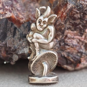 May include: A silver charm featuring a small, detailed sculpture of a goblin sitting on a mushroom. The goblin has large ears, a mischievous expression, and is holding its legs.