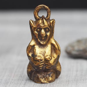 May include: A small, gold-toned metal pendant shaped like a seated gargoyle with pointed ears, a wide nose, and a circular loop at the top for hanging. The figure has a weathered, textured finish with dark highlights in the crevices of the metal.