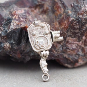 May include: A silver charm in the shape of a vintage movie camera. The charm has a detailed design with a lens, handle, and a crank.