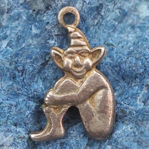 May include: A silver-toned charm in the shape of a seated goblin, with a pointed hat and smiling face. The goblin is hugging its knees. The charm has a loop at the top for attaching to a necklace or bracelet.