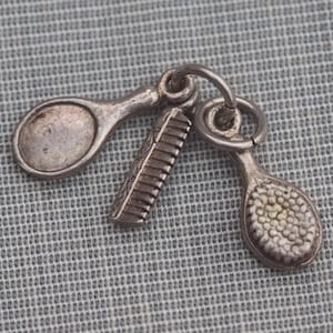 May include: A silver charm featuring a comb and two tennis racket-shaped charms. The charms are connected by a ring. The background is a gray, textured fabric.