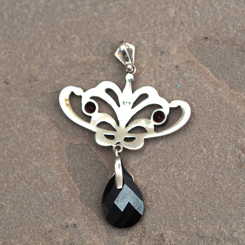 May include: Silver pendant with a black gemstone drop and two smaller red gemstones. The pendant is in the shape of a stylized butterfly with intricate details.
