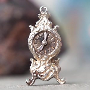 May include: A small, silver clock charm with an elaborate design. The clock face is circular with visible hands and hour markers. The charm has a decorative base and top, and is likely made of metal.