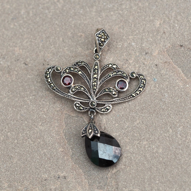 May include: A silver pendant with a black gemstone and two red gemstones. The pendant is made of intricate filigree work and has a delicate design.