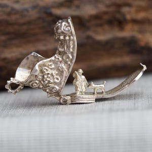 May include: A silver-colored metal hinged trinket box that opens to reveal a miniature three-dimensional scene inside. The scene features a small figure of a person carrying a pack and a small dog standing on the flat base of the open container.