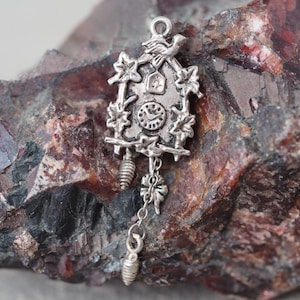 May include: A silver-colored cuckoo clock charm with intricate details. The charm features a bird perched atop the clock, a clock face, and decorative leaves. Two pinecone-shaped weights hang below the clock, connected by a chain.