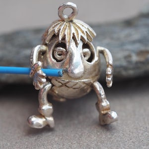 May include: A silver-toned charm in the shape of a whimsical creature with a large nose, wide eyes, and spiky hair. The charm is holding a blue stick. The creature has small arms and legs, and a loop at the top for attaching to a chain or bracelet.