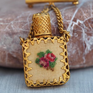 May include: A vintage gold-coloured perfume bottle pendant. The rectangular bottle features a cross-stitched floral design in pink and green on a beige background. The bottle has a decorative gold frame and a matching chain.