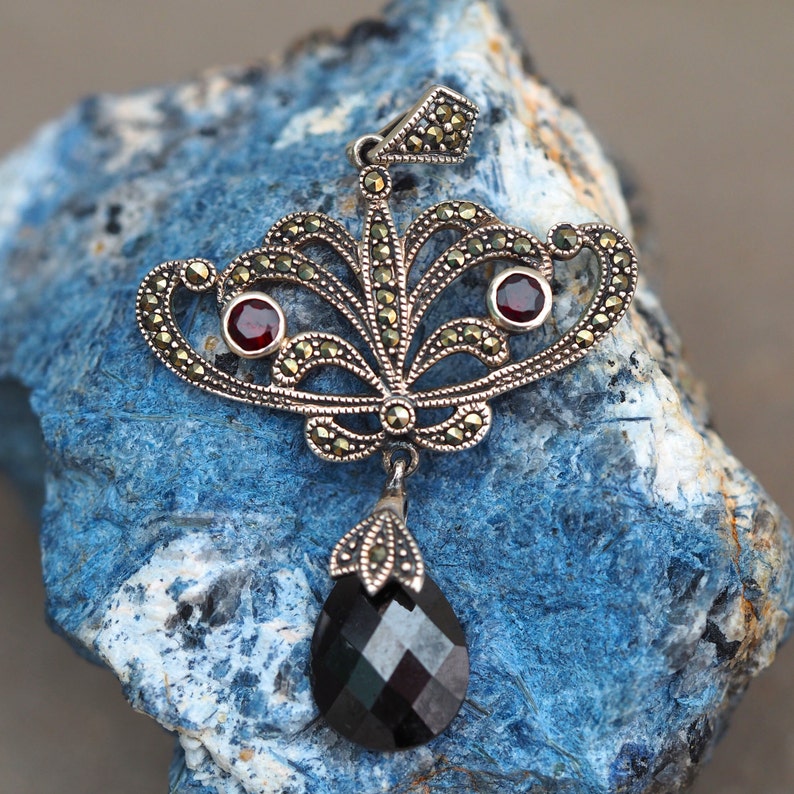 May include: A silver pendant with a black gemstone drop and two red gemstones. The pendant is made of intricate filigree work and is set on a blue background.