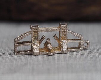 May include: A silver-toned metal charm shaped like Tower Bridge in London, featuring two vertical towers connected by a top span, with a small boat positioned between the open bascules and a circular jump ring attached to the right side.