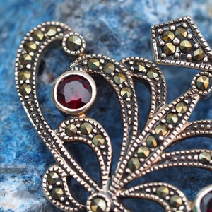 May include: A silver pendant with a delicate, intricate design featuring a central red gemstone surrounded by smaller, black gemstones. The pendant is crafted in a floral motif with a detailed, ornate style.