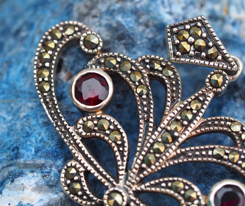 May include: A silver pendant with a delicate, intricate design featuring a central red gemstone surrounded by smaller, black gemstones. The pendant is crafted in a floral motif with a detailed, ornate style.