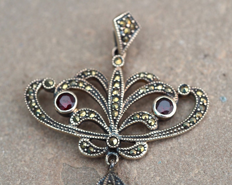 May include: A silver pendant with a delicate, ornate design featuring two red gemstones and a marcasite finish.