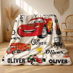 May include: A white fleece blanket featuring a red Lightning McQueen race car, a red semi-truck, a green race car, a gold trophy, and black tire graphics. The name Oliver is printed in various black fonts throughout the patterned design.