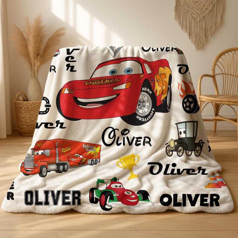 May include: A white fleece blanket featuring a red Lightning McQueen race car, a red semi-truck, a green race car, a gold trophy, and black tire graphics. The name Oliver is printed in various black fonts throughout the patterned design.
