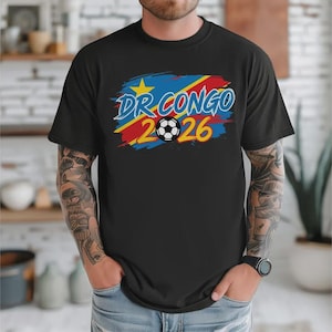 May include: Black t-shirt with a graphic design featuring the text "DR CONGO 2026" in blue and yellow, a soccer ball, and the colors of the Democratic Republic of Congo flag. The shirt is short-sleeved.