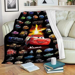 May include: A black fleece blanket featuring a repeating pattern of various Cars characters, a large central red Lightning McQueen graphic with the number 95, and the word Name in white script. The blanket has a white plush underside.