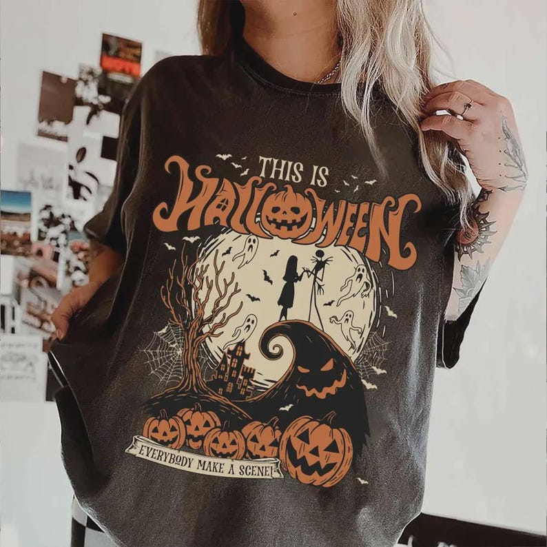 K&ouml;nnte beinhalten: Dunkelgraues T-Shirt mit einem Halloween-Motiv. Das Design zeigt den Text "THIS IS HALLOWEEN" in Orange, zusammen mit K&uuml;rbissen, Geistern und einer gruseligen Szene. Der Satz "EVERYBODY MAKE A SCENE!" ist ebenfalls enthalten.