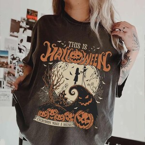K&ouml;nnte beinhalten: Dunkelgraues T-Shirt mit einem Halloween-Motiv. Das Design zeigt den Text "THIS IS HALLOWEEN" &uuml;ber einer gruseligen Szene mit Vollmond, Jack Skellington und K&uuml;rbissen. Der Satz "EVERYBODY MAKE A SCENE!" ist darunter gedruckt.