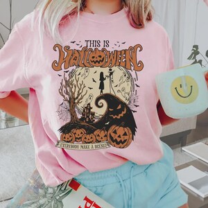 K&ouml;nnte beinhalten: Hellrosa Halloween-T-Shirt mit einem grafischen Design. Das Design zeigt einen K&uuml;rbis, Geister, einen Hexenhut und den Text "THIS IS HALLOWEEN" und "EVERYBODY MAKE A SCENE!".