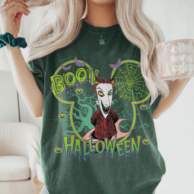 May include: Dark green t-shirt featuring a Halloween-themed design. The graphic includes the word "BOO!!" in green, a character with a white face and red hair, and the word "HALLOWEEN" in green. The design is set within a Mickey Mouse-shaped outline.