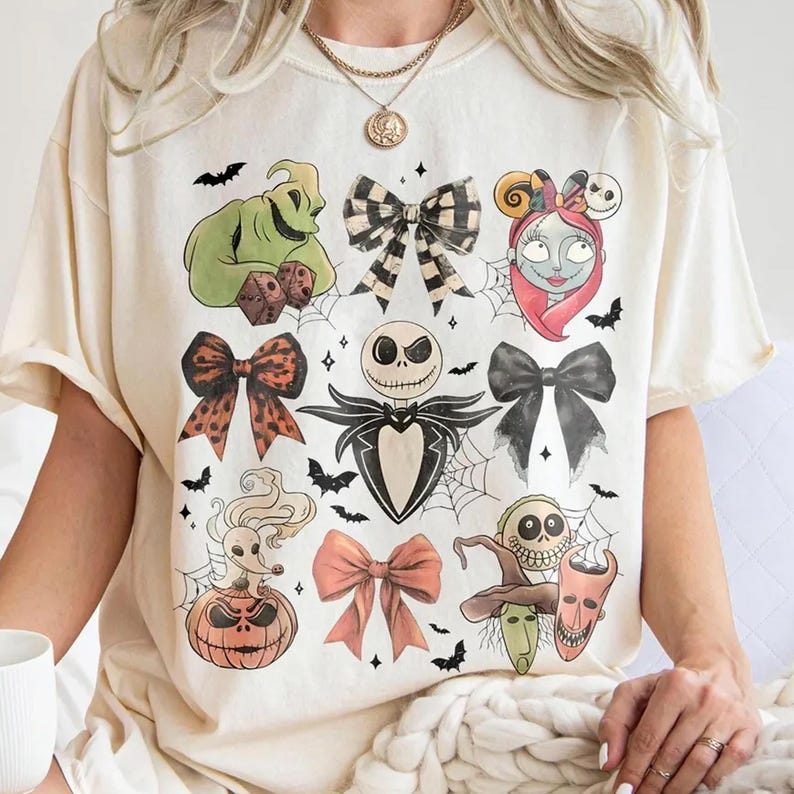 Może przedstawiać: Kremowa koszulka z kolorowymi ilustracjami postaci i kokard z filmu "Miasteczko Halloween". Projekt zawiera Jacka Skellingtona, Sally, Oogie Boogie i inne elementy związane z Halloween. Zawiera r&oacute;wnież nietoperze i pajęczyny.