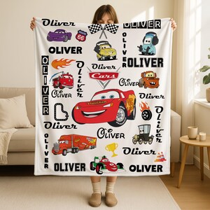 May include: A white fleece blanket featuring a repeating pattern of characters and icons from the Cars movie franchise, including Lightning McQueen, Mater, and a Route 66 sign. The name Oliver is printed in various fonts throughout the design.