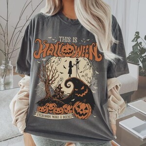 K&ouml;nnte beinhalten: Dunkelgraues T-Shirt mit einem Halloween-Motiv. Das Design zeigt den Text "THIS IS HALLOWEEN" &uuml;ber einer Vollmondszene mit Jack Skellington, K&uuml;rbissen, Geistern und einem Spukhaus. Der Satz "EVERYBODY MAKE A SCENE!" steht unten.