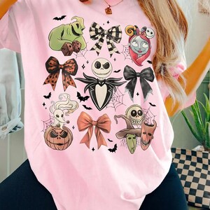 Może przedstawiać: R&oacute;żowa koszulka z kolażem postaci z Halloween i wzorami kokard. Grafika zawiera obrazy Jacka Skellingtona, Sally, Oogie Boogie i innych postaci z filmu. Projekt zawiera r&oacute;wnież pajęczyny i nietoperze.