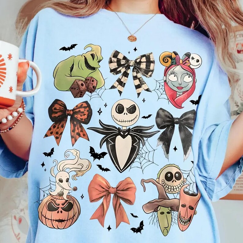 Może przedstawiać: Jasnoniebieska koszulka z ilustracjami o tematyce Halloween. Projekt zawiera postacie takie jak Jack Skellington, Sally i Oogie Boogie, a także kokardy, nietoperze i pajęczyny. Wygodny, codzienny element garderoby.