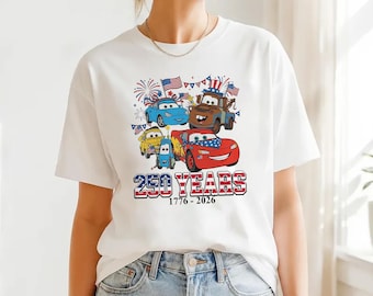 May include: White t-shirt with a graphic of cartoon cars decorated with American flags and fireworks. The text "250 YEARS 1776-2026" is in red, white, and blue. The shirt has a crew neck and short sleeves.