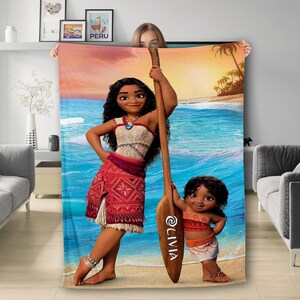 Puede incluir: Una manta de forro polar rectangular con el estampado de una joven y un ni&ntilde;o peque&ntilde;o en una playa junto a un gran remo de madera. El nombre LIVIA est&aacute; impreso verticalmente en blanco sobre el remo. El fondo muestra olas azules.