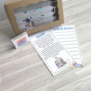 May include: A brown cardboard box with a window containing white candles, a small matchbox with a colorful logo, and two printed cards. One card is titled ORACIÓN and the other LISTA DE DESEOS, both featuring illustrations and text in Spanish.