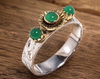 May include: A silver ring featuring a hammered band and a decorative top element with three round green cabochon stones set in gold-toned metal. The central stone is larger and surrounded by a scalloped gold-toned petal-like frame design.
