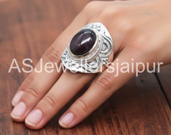 Könnte beinhalten: Ein großer, verzierter Silberring mit einem dunkelvioletten, ovalen Edelstein. Der Ring hat aufwendige Details um den Stein herum und ein breites Band. Der Text "ASJewelsjaipur" ist sichtbar.